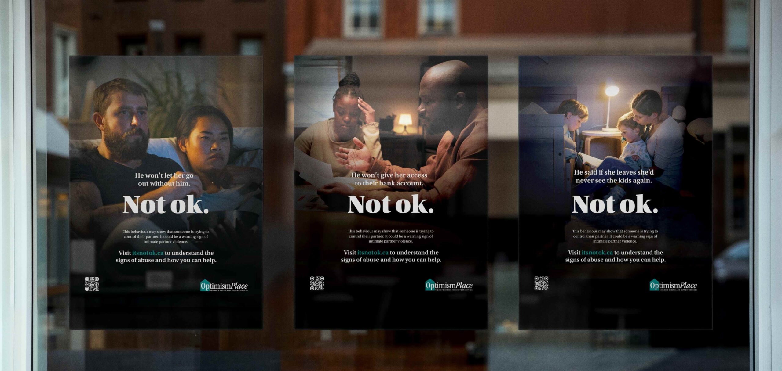 Three Optimism Place Not ok. campaign posters displayed in a window, each showing a different household or relationship scenario with the campaign headline.