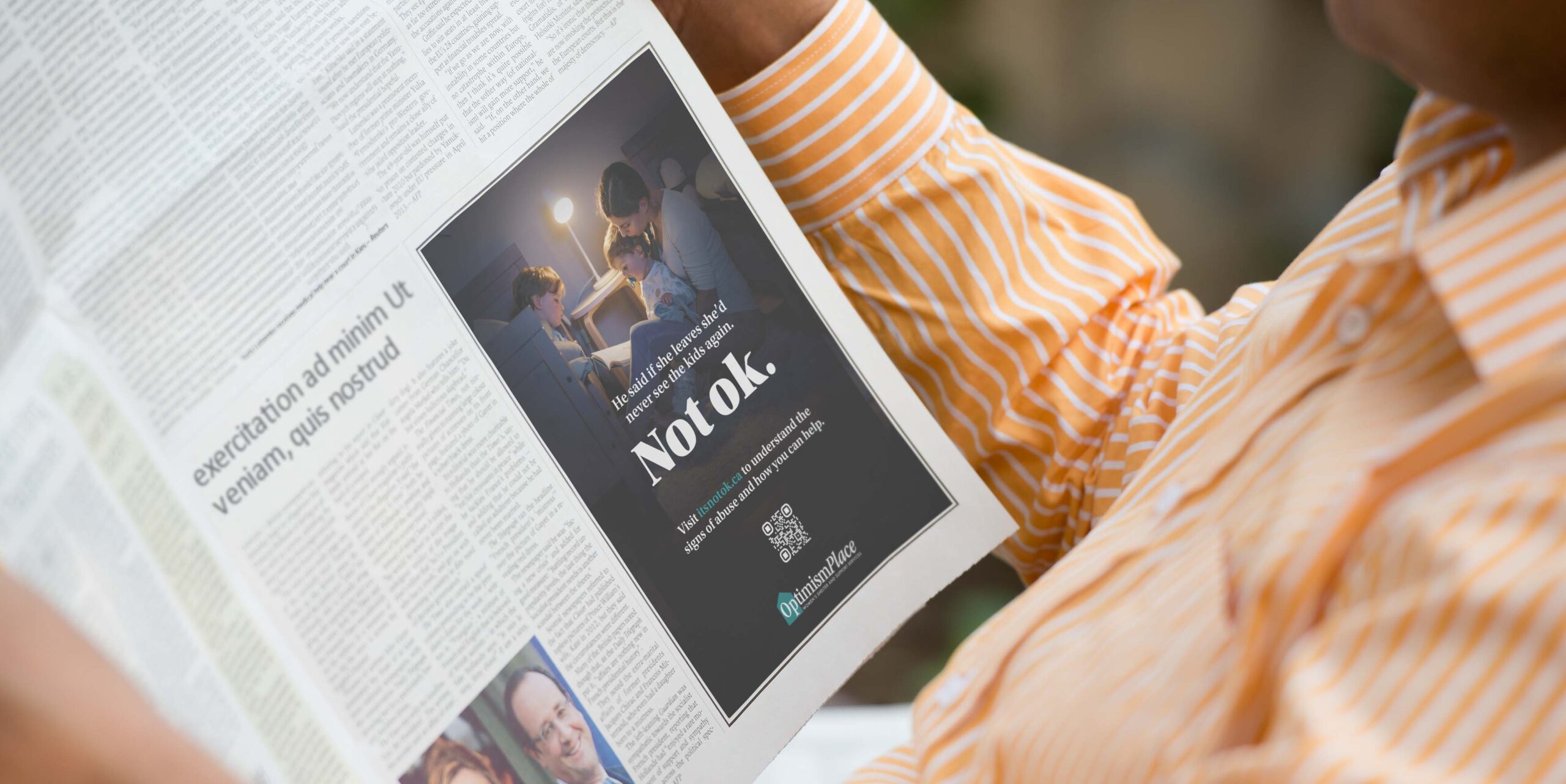 Newspaper open to a full-page Not ok. campaign ad for Optimism Place showing a parent reading to two children, with the campaign headline and QR code visible.