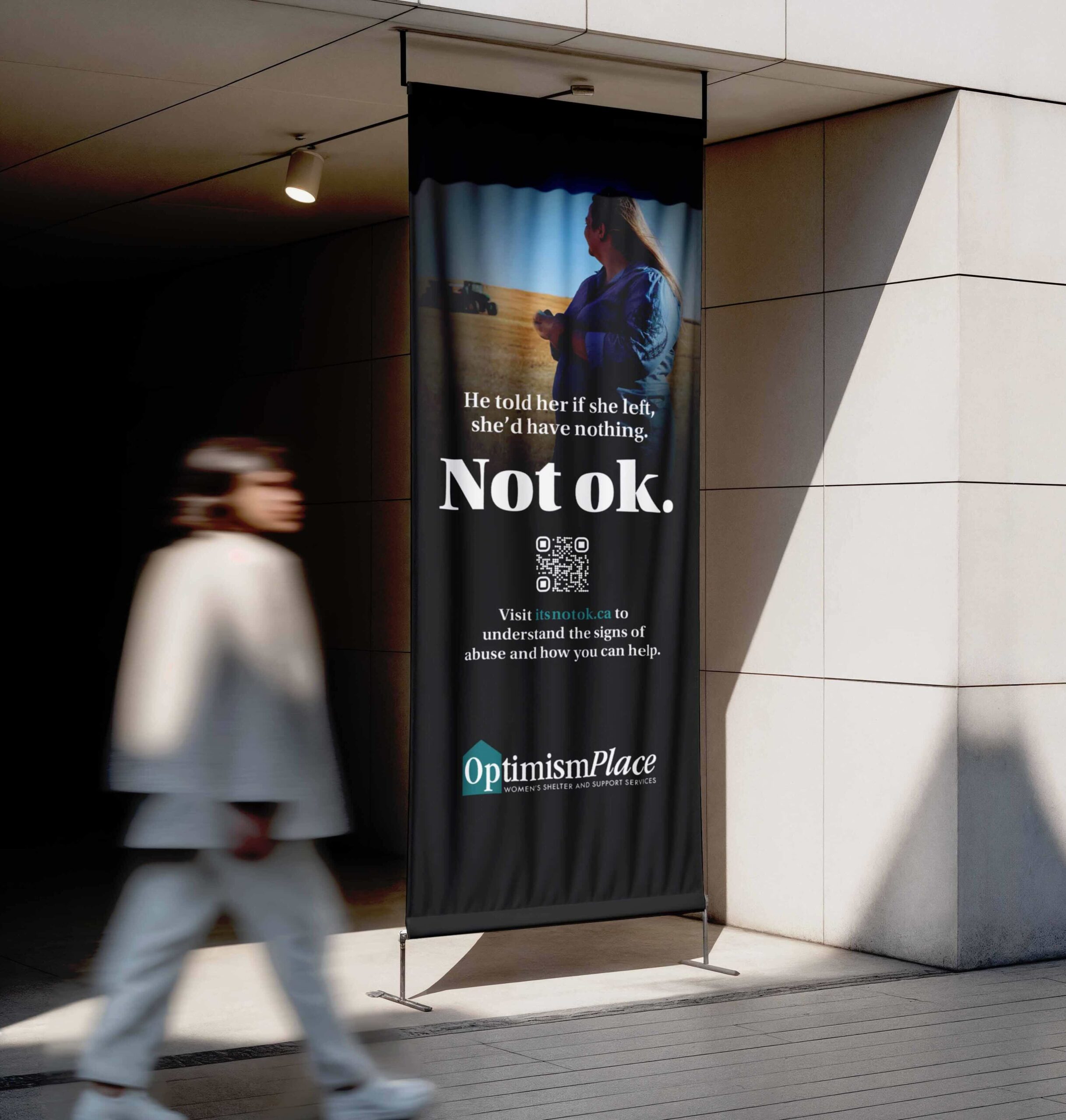 Vertical outdoor banner for the Optimism Place Not ok. campaign showing a woman standing in a rural setting with the campaign headline and QR code.